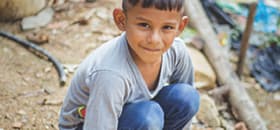A boy crouches on the ground and smiles up at the camera