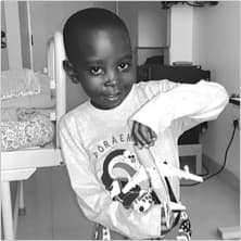 Saviour smiles and plays with toy plane. He's standing next to his hospital bed.