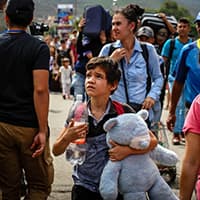 A boy walks across a bridge with a crowd of refugees. He holds a big teddy bear in his arms and looks nervous