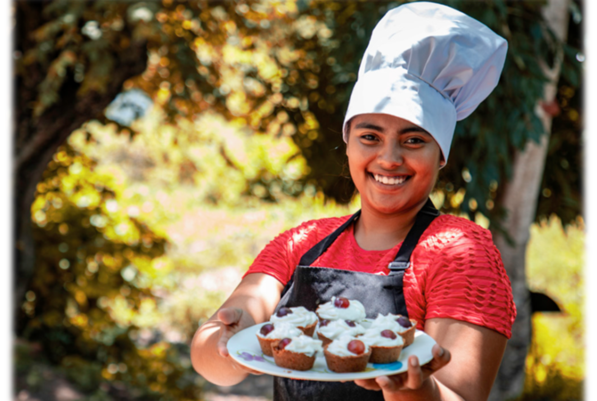 A smiling young woman wearing a chef's hat is holding out a plate of frosted bakery items.