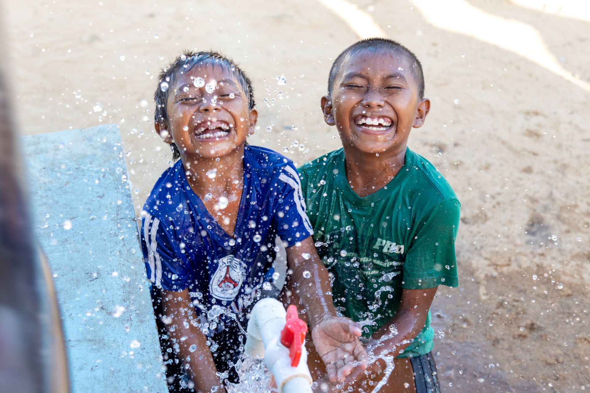 Two laughing boys play in clean water from a spout outdoors.