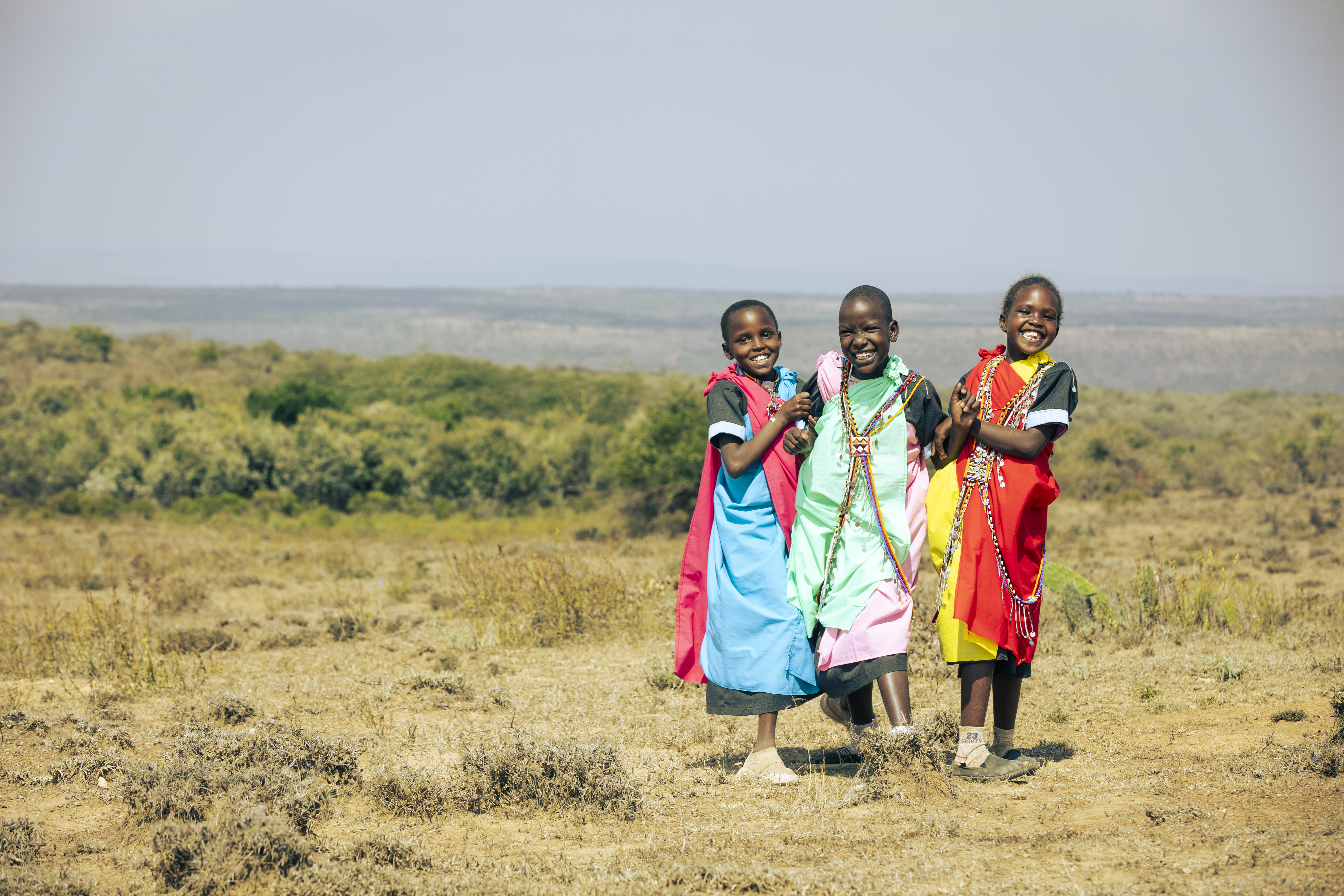 Three Kenyan girls wearing brightly coloured clothing are embracing each other on top of a hill with the horizon view behind them and are smiling into the camera.