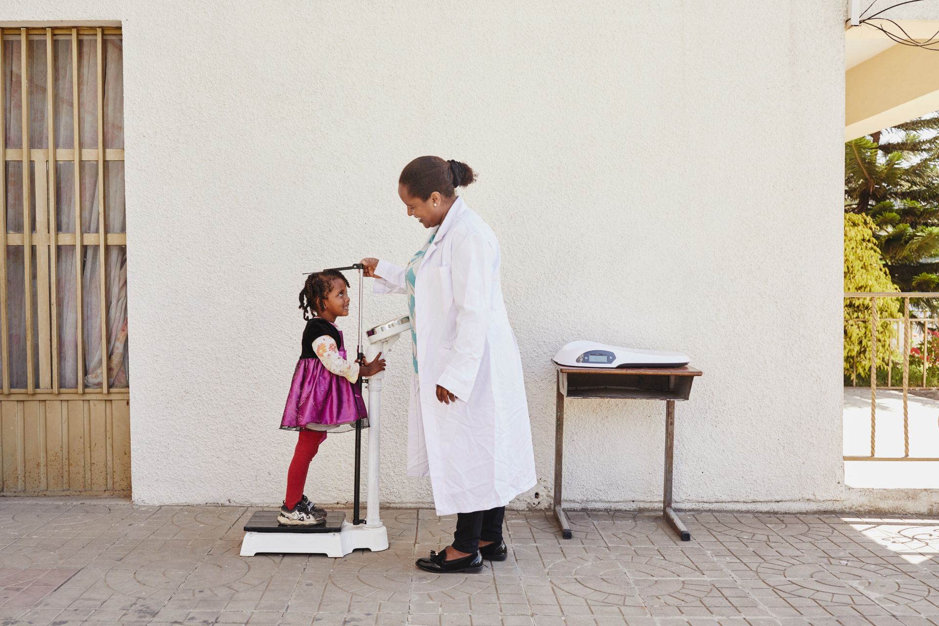 A little girl is smiling up at a female doctor as she has her height measured outdoors.