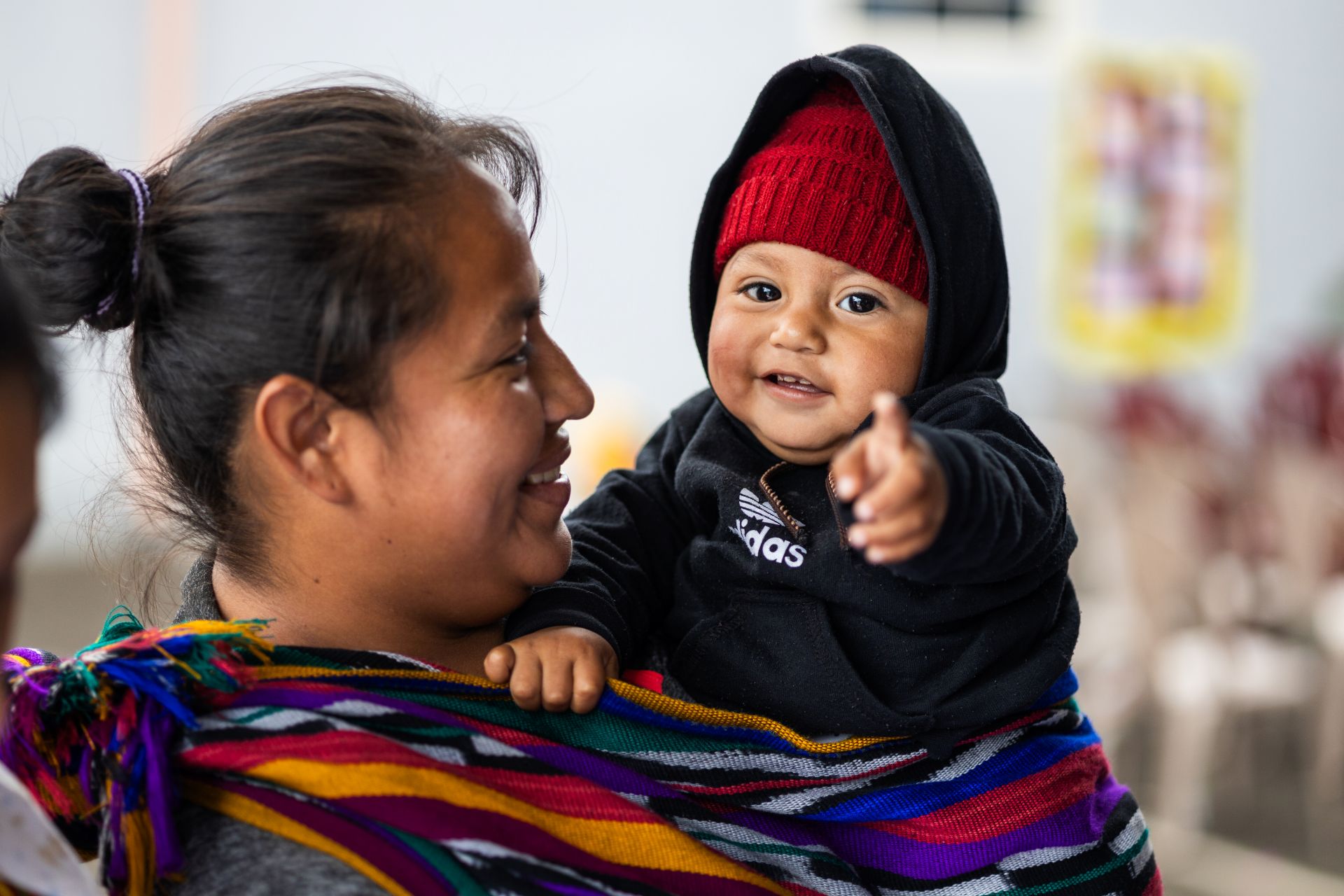 Smiling mom holds her toddler in her arms, who is pointing at the camera.