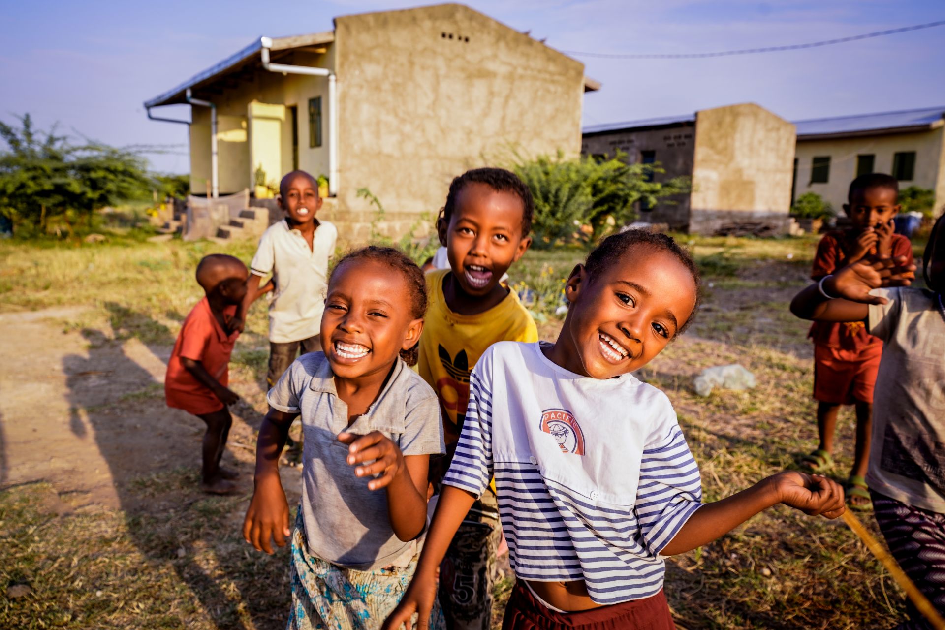 A group of children are smiling into the camera while playing outside with buildings in the background.