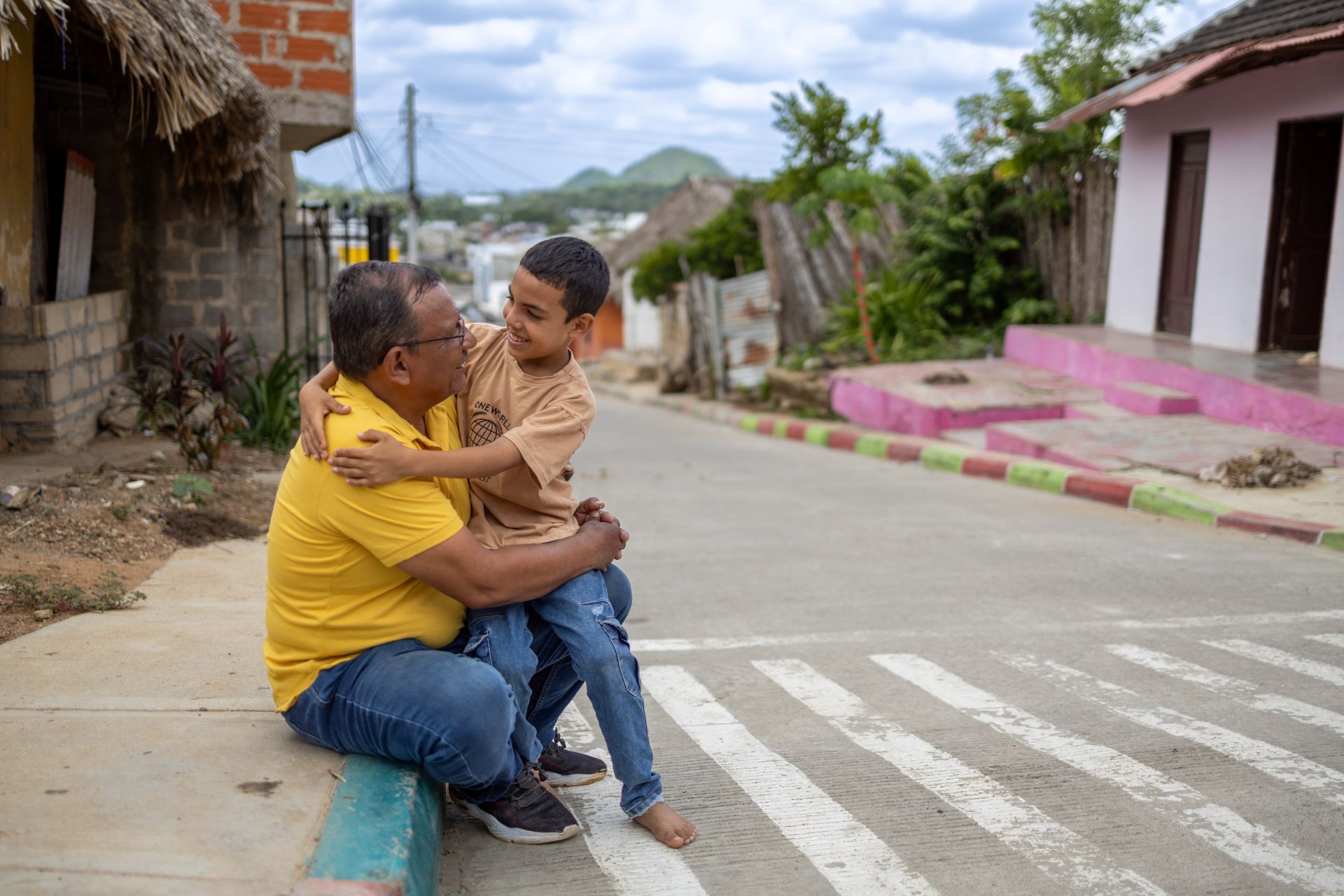 A dad sits on a sidewalk in his community holding his smiling son in his arms.
