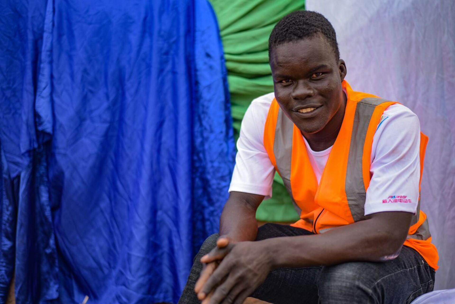 Nourou is sitting down outside, wearing a white shirt and an orange vest. There is a blue piece of fabric behind him.