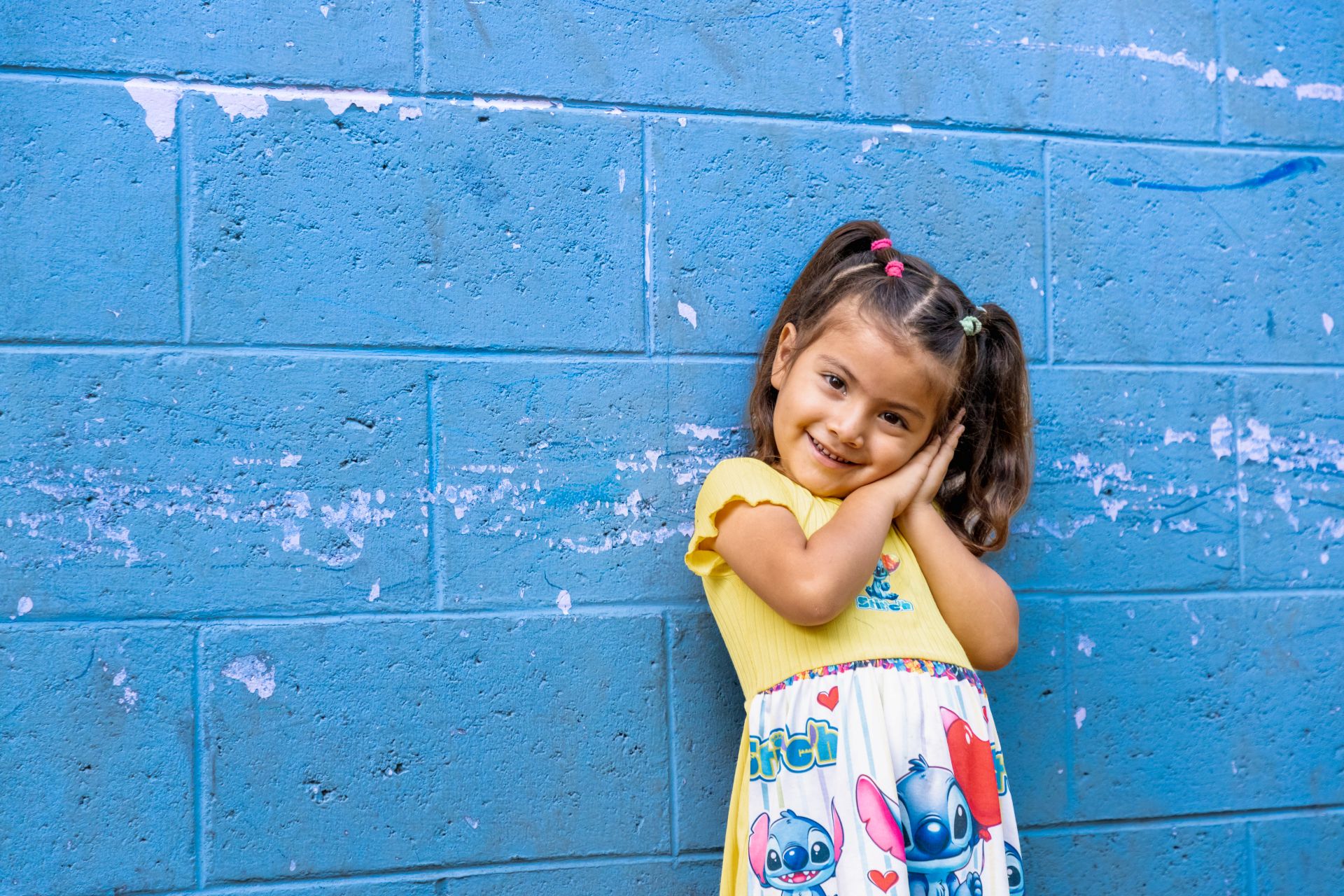 A young girl wearing a colourful dress and standing in front of a painted blue wall, smiles into the camera.