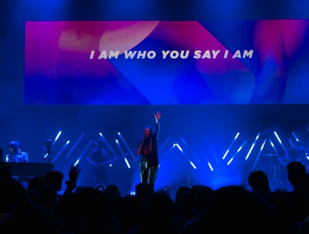 Brooke Nicholls leads worship on a large stage. There's a large projection screen behind her with the words, I am who you say I am, on it.