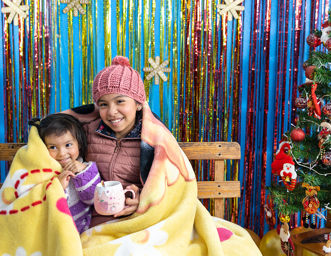 Camila sits with her sister in a sparkly christmas sceen. There are streamers in the background and a christmas tree beside them. They are wrapped in a warm, yellow blanket.