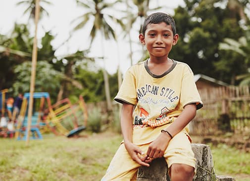 A boy in a t-shirt sits on a bench and smiles at the camera. /></div>
<script src=