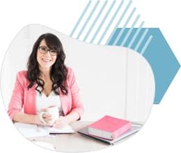 A woman in a pink jacket sits at her desk with a cup of coffe and some books. She smiles.