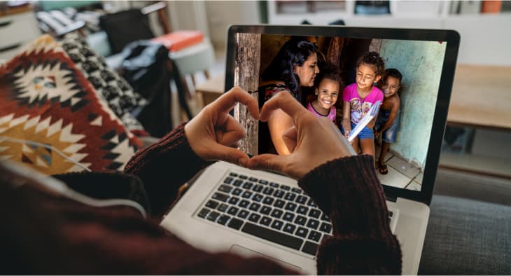 A woman sits in front of a laptop and makes a heart shape with her hands. On screen, a mom and three children smile back.