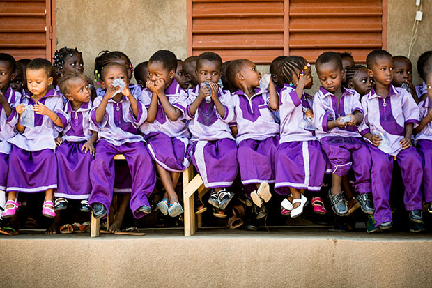 A large group of children sit lined up on benches outside a compassion centre. They are all wearing purple uniforms and having a snack.