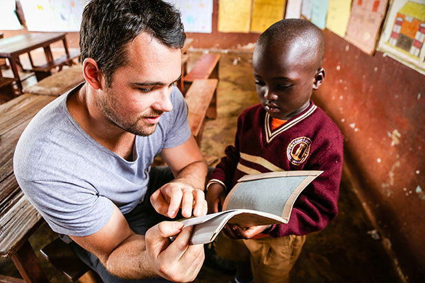 A young man sits in at a table and reads a book with a young boy.